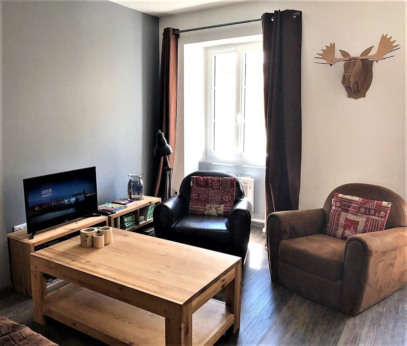 Un salon avec un téléviseur, une table en bois, deux chaises, une fenêtre avec des rideaux et une tête de cerf empaillée accrochée au mur.