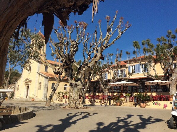 A tree-lined square with a building that has a yellow facade and red-tiled roof. There are outdoor tables and chairs, and a sign that reads "Villa Sainte-Anne." - In the heart of the village, luxury studio 17m2, flowered terrace 9 m2, air conditioning (Porquerolles)