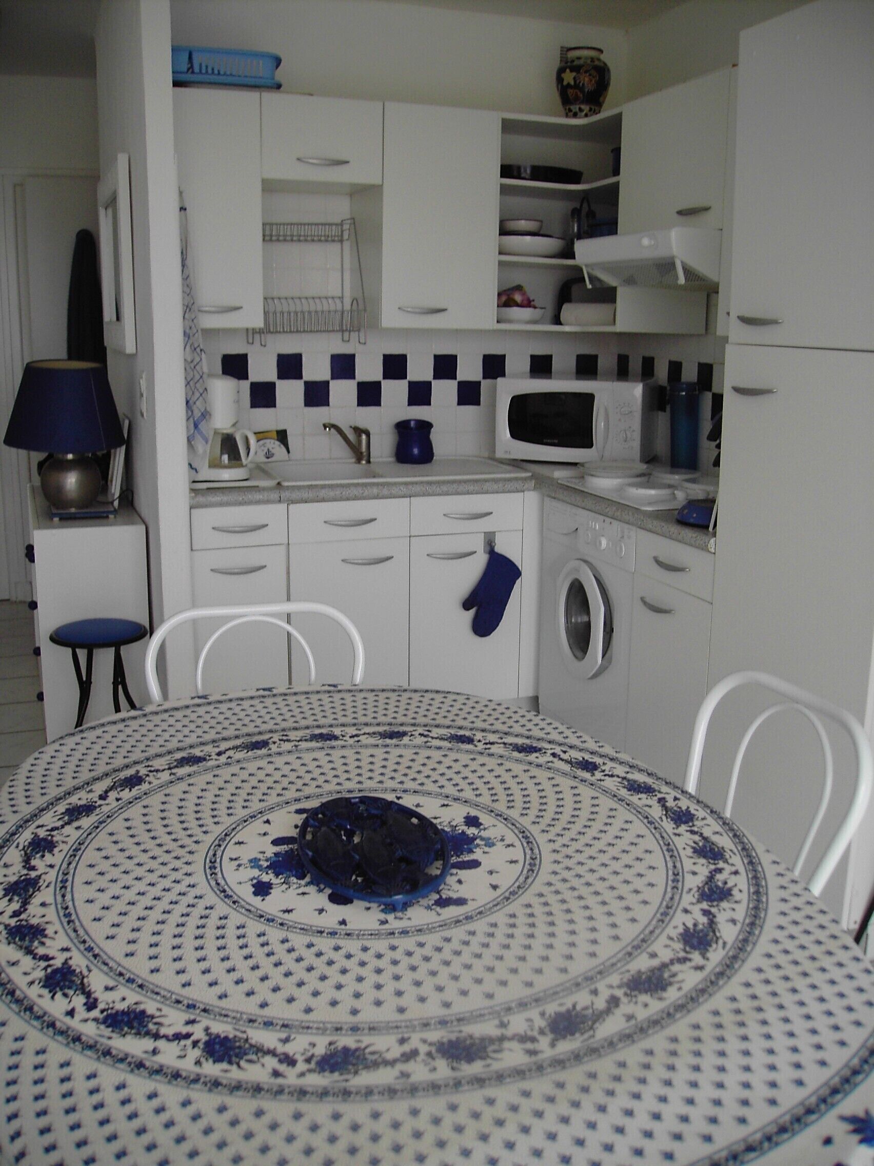 A compact kitchen with white cabinets, a microwave, a washing machine, and a dining table with a blue and white patterned tablecloth.