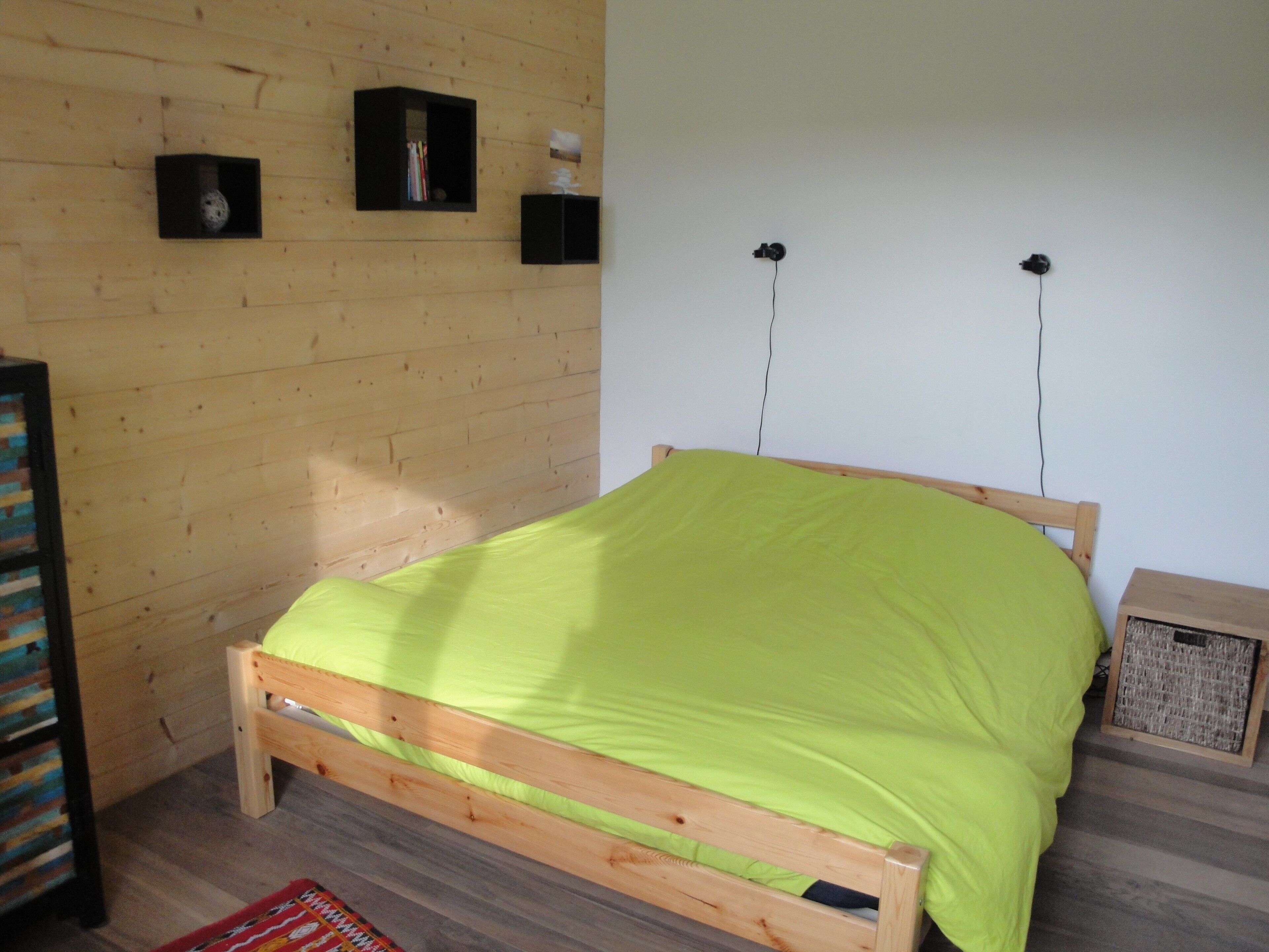 Ein hölzernes Bett mit grüner Bettdecke in einem Zimmer mit Holzwänden, einem kleinen Bücherregal und einem kleinen Lautsprecher an der Wand.