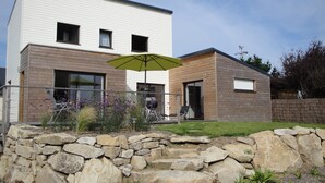 A modern two-story house with a stone retaining wall, a patio with an umbrella, and a barbecue setup.