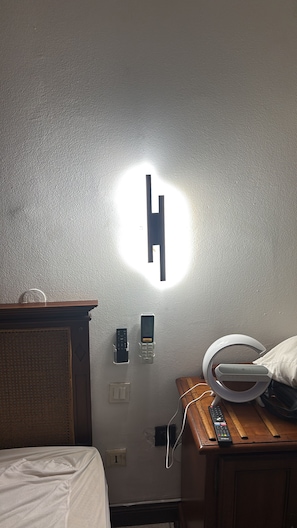 Une chambre d’hôtel comprenant un lit, une table de chevet avec une lampe, un luminaire fixé au mur et un panneau de commande.