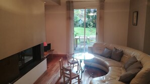 Un salon comprenant un canapé, un téléviseur, une table et des chaises. Une porte-fenêtre coulissante donne accès à un espace extérieur avec une pelouse et une table.