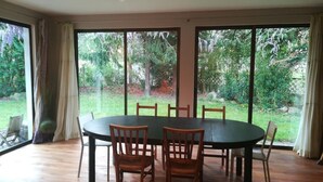 Un coin repas avec une table ronde et des chaises, de grandes fenêtres avec des rideaux et une vue sur la verdure à l’extérieur.
