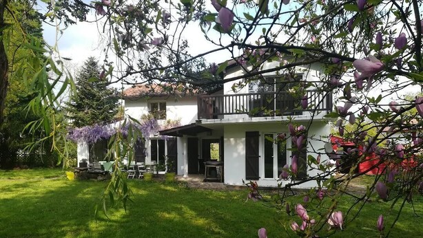 Une maison à deux étages dotée d’un balcon, entourée d’une pelouse bien entretenue et d’arbres en fleurs.