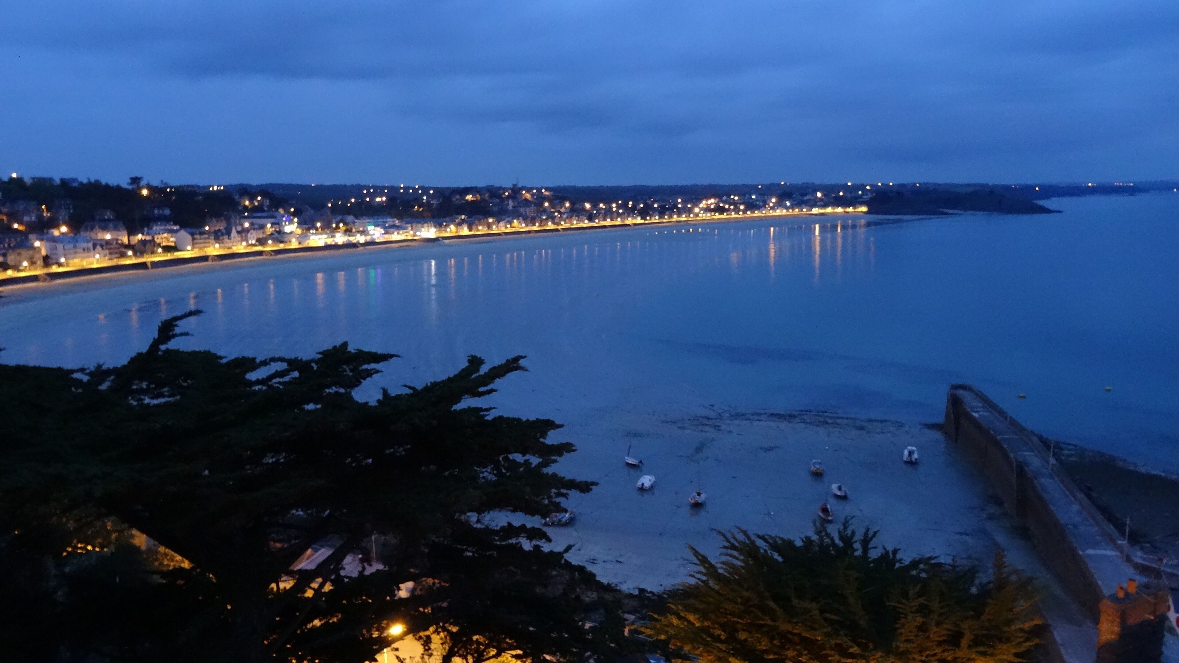 Un paisaje urbano costero al atardecer con un mar tranquilo, un paseo marítimo y luces de la ciudad a lo lejos.