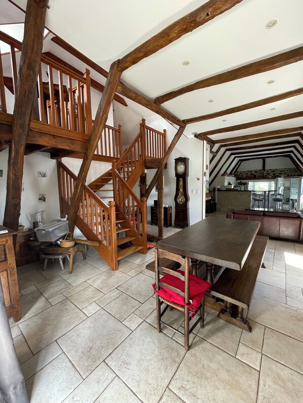A spacious room with a high ceiling, exposed wooden beams, a large dining table, and a staircase leading to an upper level.