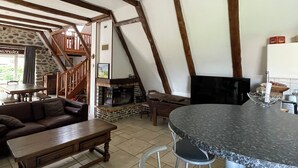 A spacious living area with exposed wooden beams, a stone fireplace, a dining table, and a kitchen area with bar stools. - Fully restored stone and slate barn in a typical village (Lacroix-Barrez)