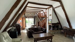 An attic living space with a sloped ceiling, wooden beams, a stone fireplace, a sofa, a dining table, and a staircase leading to an upper level. - Fully restored stone and slate barn in a typical village (Lacroix-Barrez)