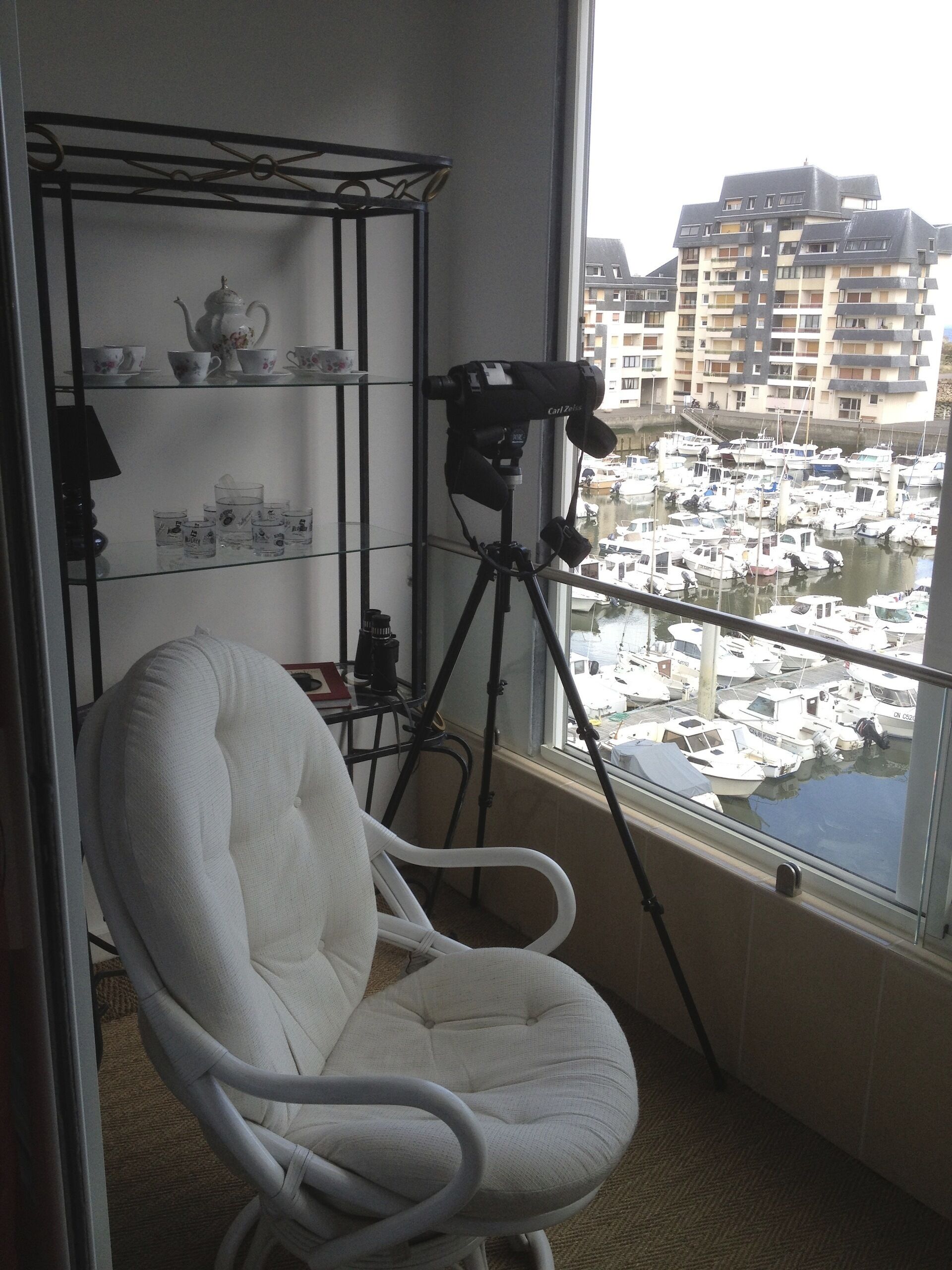 Ein Zimmer mit einem weißen Stuhl, einem Stativ mit einer Kamera und Blick auf einen Yachthafen mit Booten.