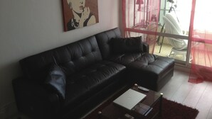 A black leather sofa with a laptop and remote control on a glass coffee table. A painting is on the wall, and a chair is visible through a glass door. - BEAUTIFUL WATERFRONT APARTMENT - BREATHTAKING MARINA AND SEA VIEWS! (Courseulles-Sur-Mer)