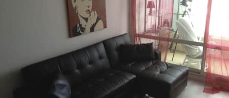 Un sofá de cuero negro con una laptop y un control remoto sobre una mesa de centro de vidrio. Un cuadro adorna la pared, y se ve una silla a través de una puerta de vidrio.