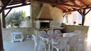 An outdoor patio with a stone wall, a pizza oven, white plastic chairs, and a table with a floral tablecloth. - Country house with heated swimming pool with solar panels. (Mauroux)