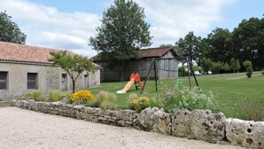A rustic stone wall with a playground, a stone building, and a wooden barn in the background. - Country house with heated swimming pool with solar panels. (Mauroux)