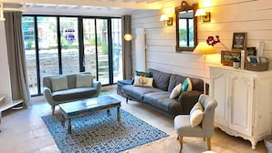 Ein Wohnzimmer mit einer Couch, einem Sessel, einem Couchtisch und einem Schrank. Ein großes Fenster mit Vorhängen, ein Spiegel und Wandlampen sind ebenfalls vorhanden.