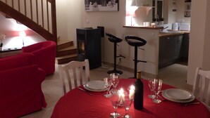 Un coin repas avec une nappe rouge, des assiettes blanches et des verres à vin. On y trouve deux tabourets de bar noirs et une chaise en bois. Un escalier est visible au fond, menant à une autre pièce.