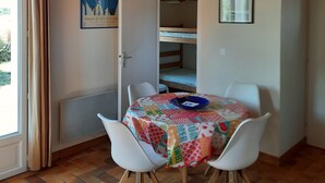 Een kamer met een houten plafond, een tafel met een kleurrijke tafelkleed, witte stoelen, een stapelbed en een raam met gordijnen.