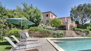 Un espace piscine avec des chaises longues et un parasol, un mur en pierre et une maison à deux étages dotée d’un balcon.