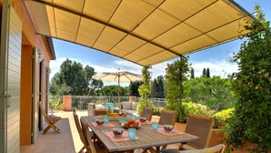 Une terrasse extérieure avec une table dressée pour le repas, entourée de chaises, et un grand parasol offrant de l’ombre.