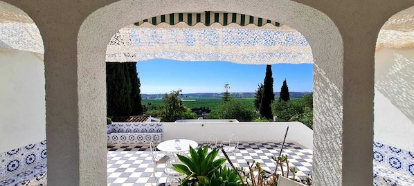 A terrace with a checkered floor, a patterned bench, and a view of a green landscape with trees.