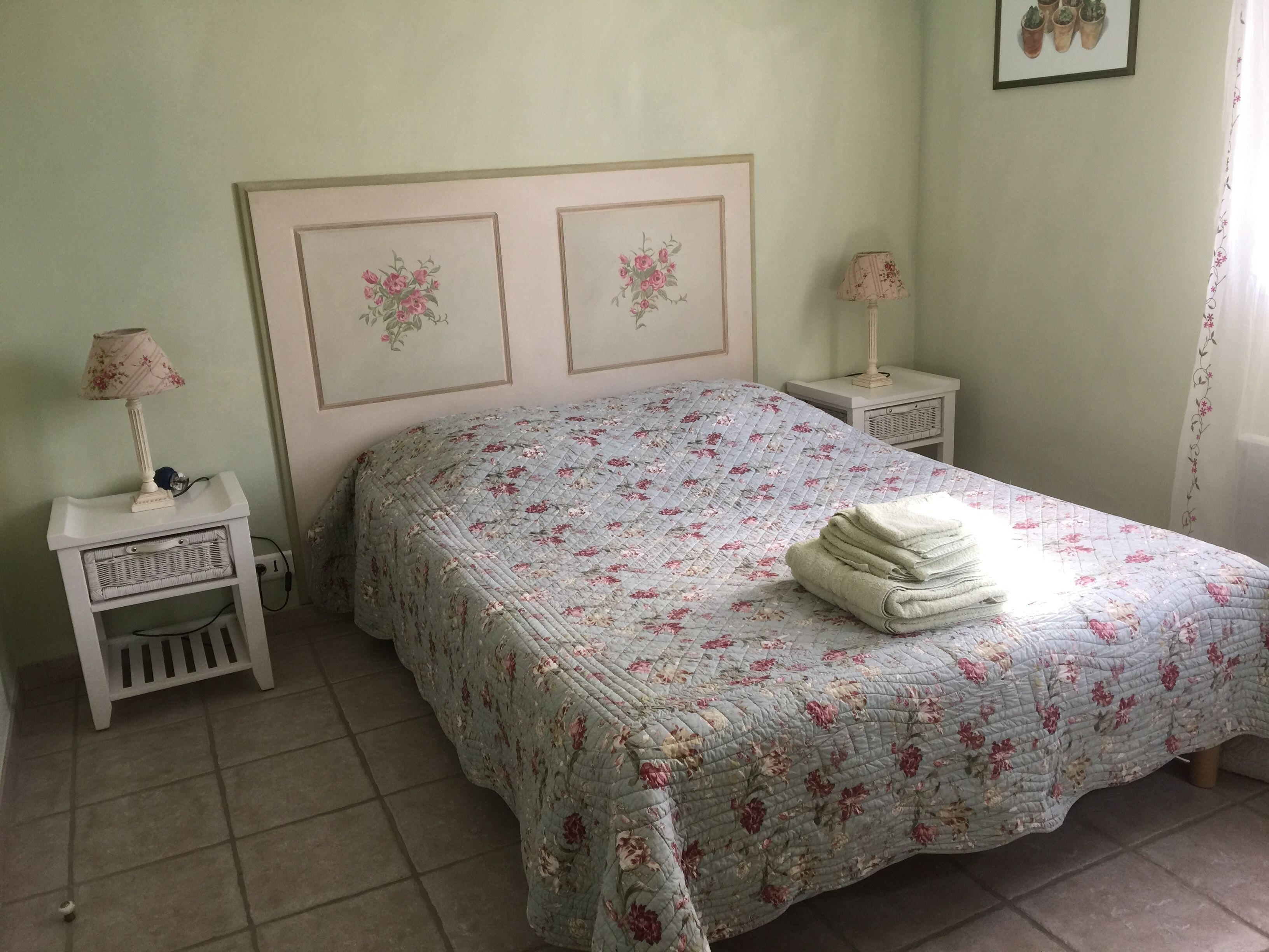 Ein Schlafzimmer mit floralem Bettbezug, zwei Nachttische mit Lampen, einem Weidenbeistelltisch und einem gerahmten Blumenbild an der Wand.