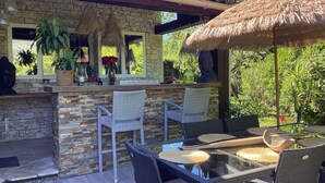 Zona de bar al aire libre con paredes de piedra, sillas de mimbre y un toldo de paja.
