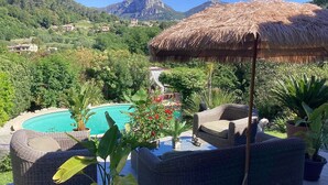 Zona de piscina con mobiliario de mimbre, una sombrilla de paja y vista a una montaña y un pueblo al fondo.