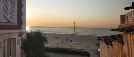 Vista de la playa con un edificio en primer plano, cielo despejado y el sol poniéndose sobre el agua.