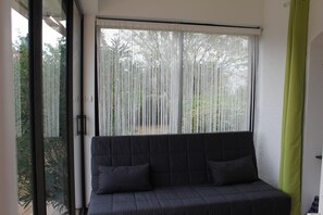 A room with a dark-colored sofa, a large window with sheer curtains, and a green curtain on the right.