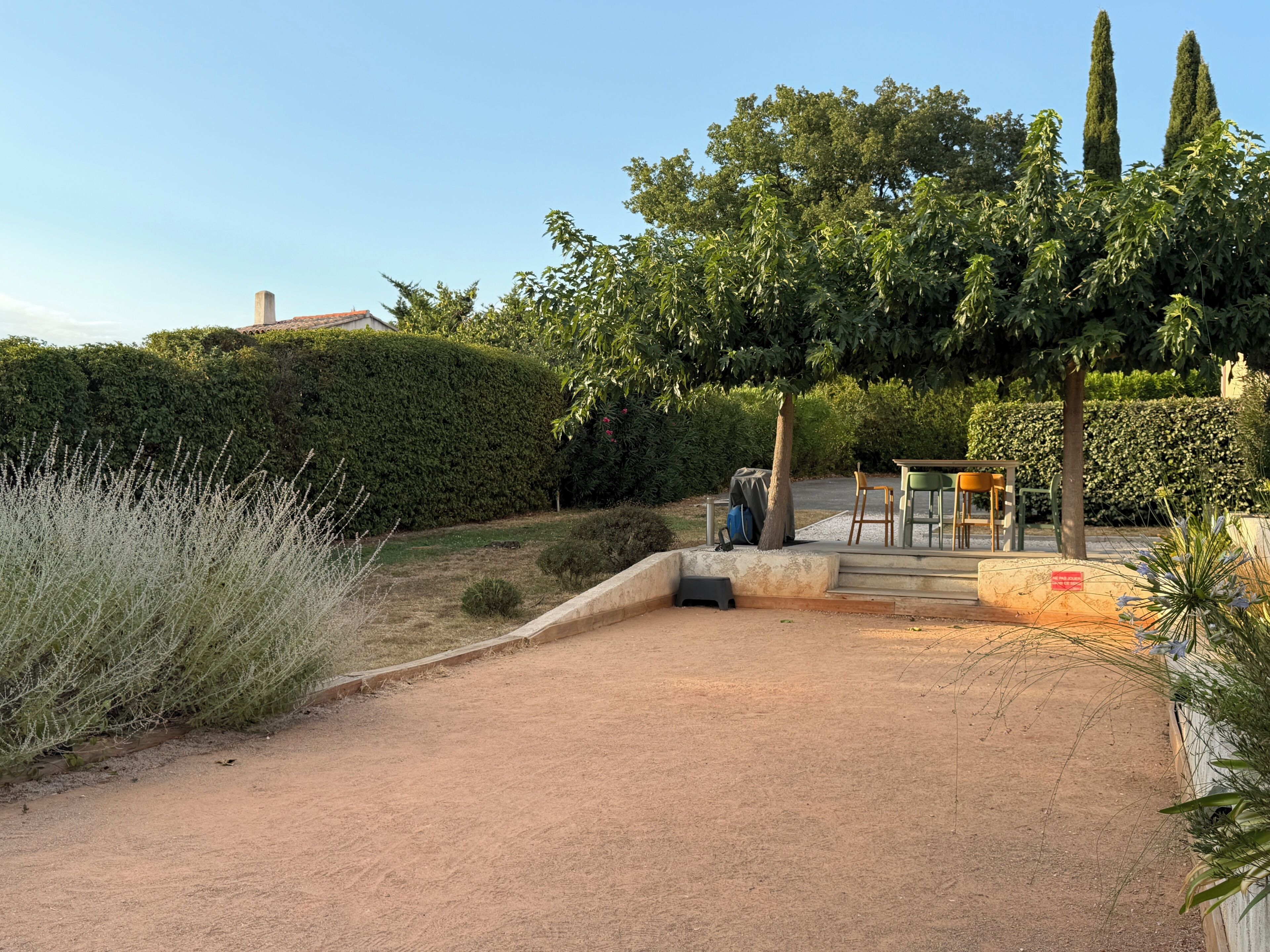 A paved pathway leading to a seating area with a table and chairs, surrounded by greenery and a hedge.