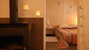 A room with a wood-burning stove, a bed with patterned bedding, and a nightstand with lamps. - Adorable guest apartment at the pool (Prayssas)