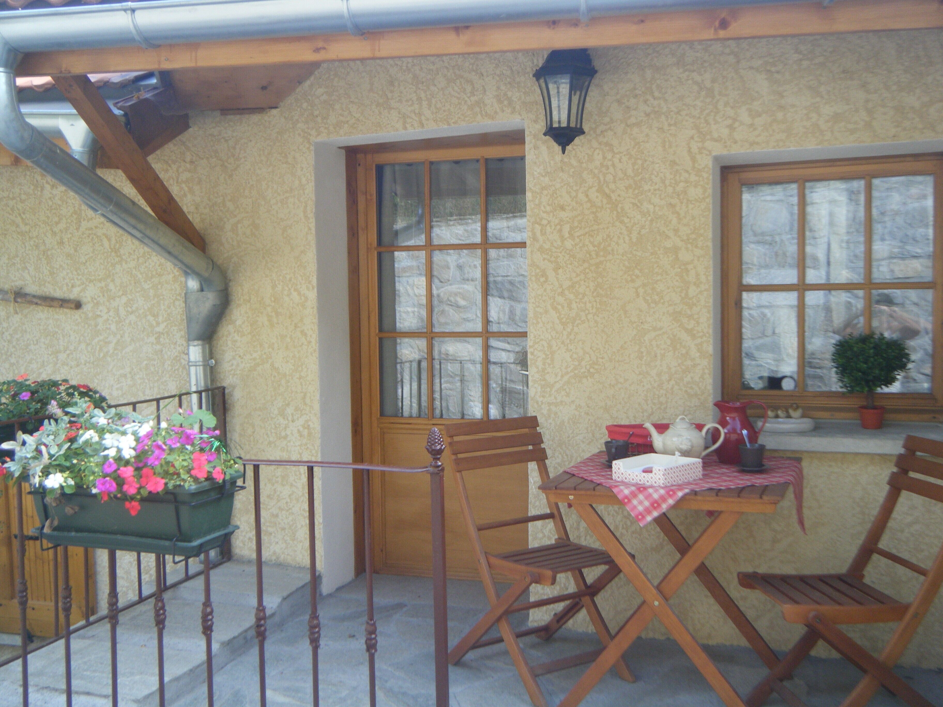 A cozy outdoor seating area with a wooden table and chairs, a flower planter, and a window with a view.