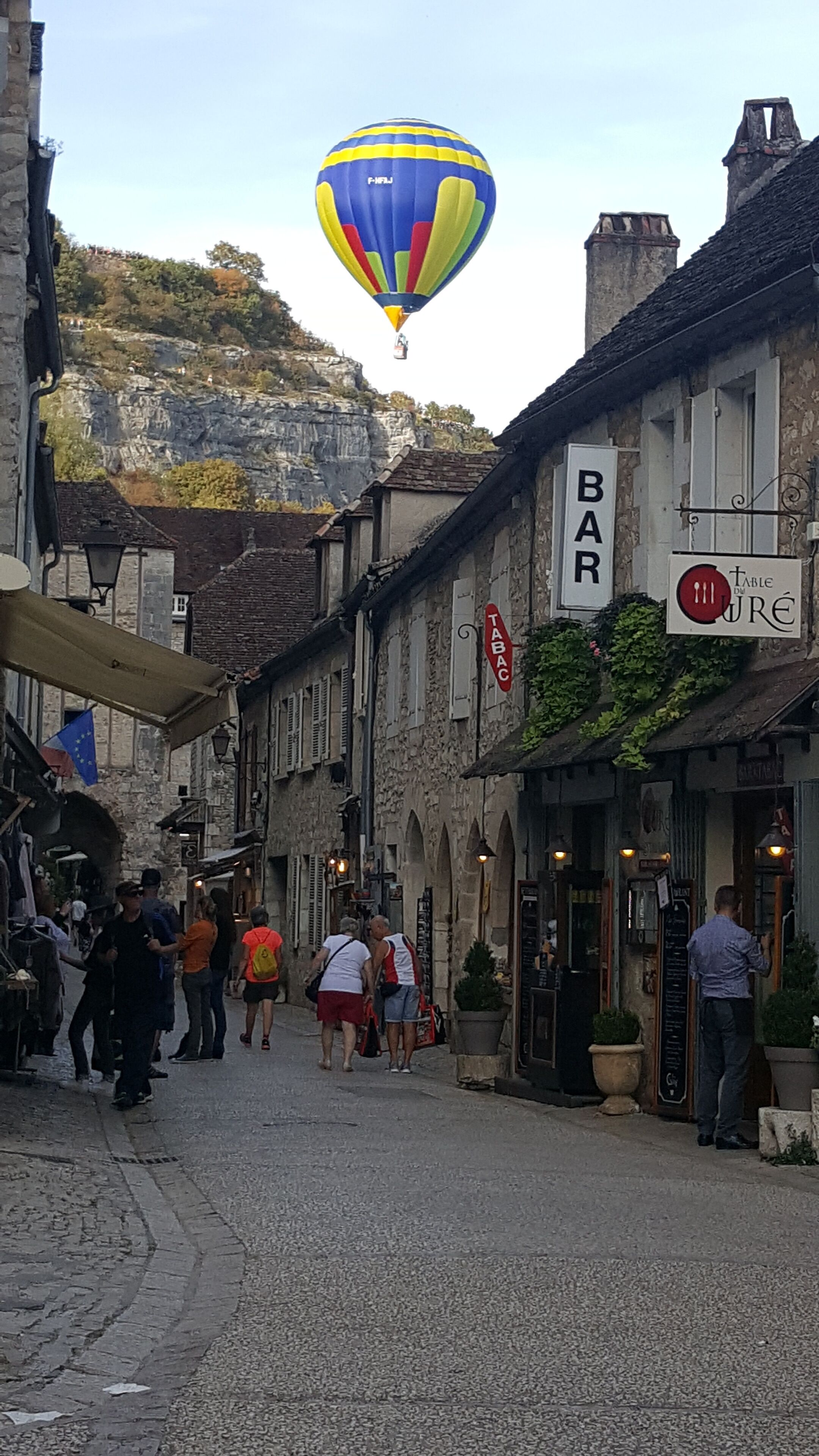 A narrow street with buildings on either side, a bar sign, and a hot air balloon in the background.