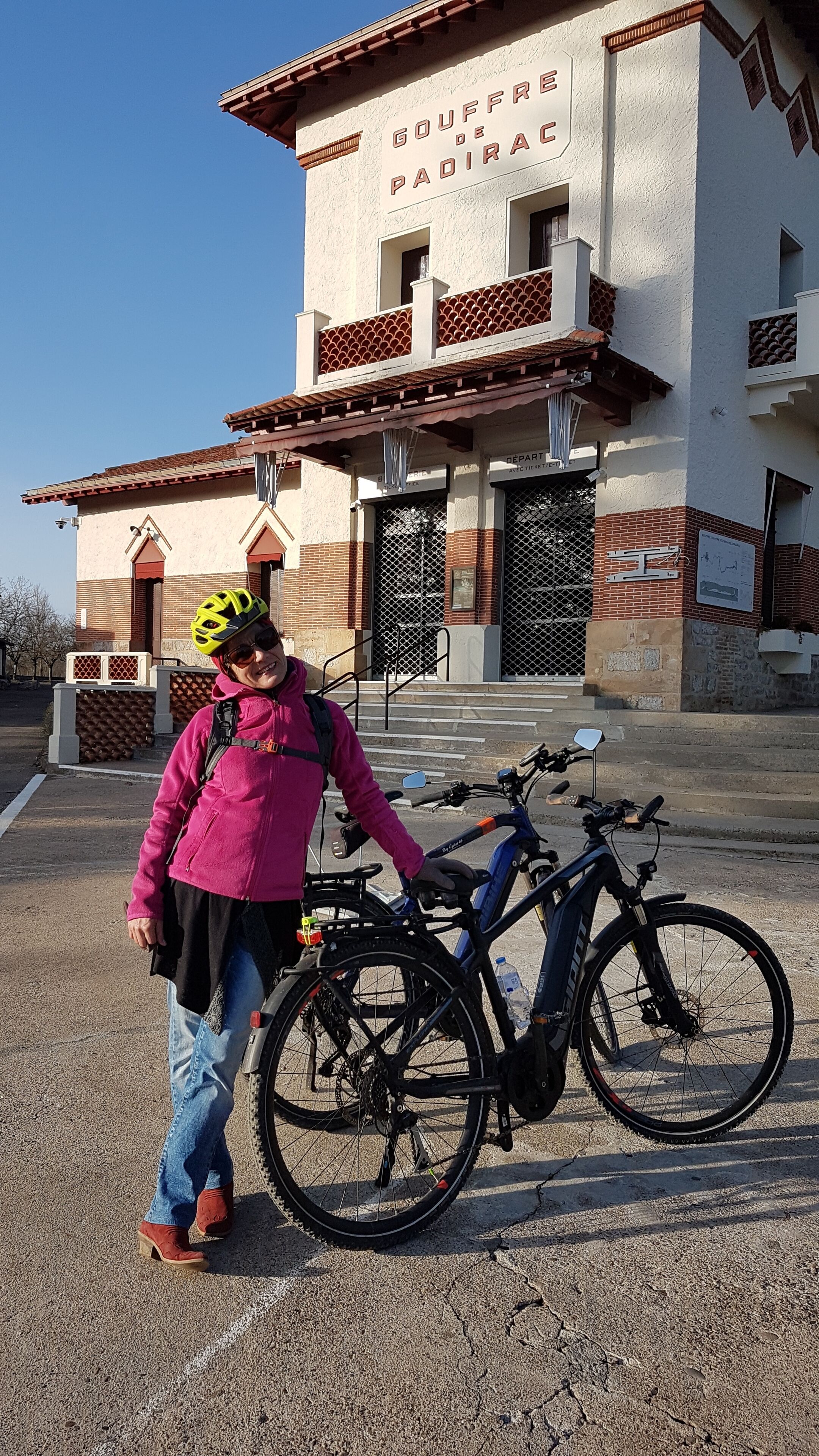 A person standing next to a bicycle in front of a building with a sign that reads 'GOUFFRE DE PADIRAC.'
