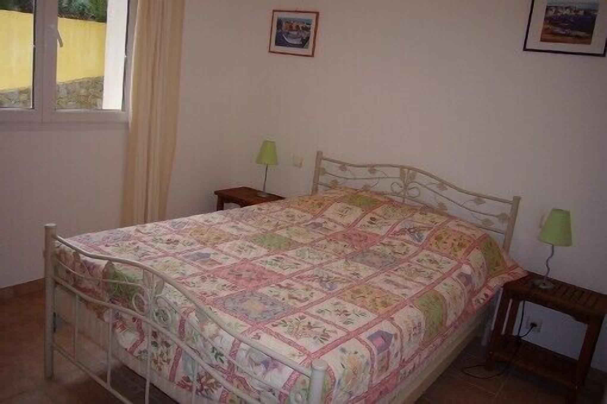 Une chambre à coucher avec un lit en métal, une couvre-lit matelassé, deux tables de chevet avec des lampes et une fenêtre avec des rideaux.