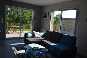 A modern living room with a sectional sofa, a glass coffee table, and a sliding glass door leading to a patio area. - The closeau du cèdre in Normandy between Paris and Rouen, near Giverny (Ménilles)