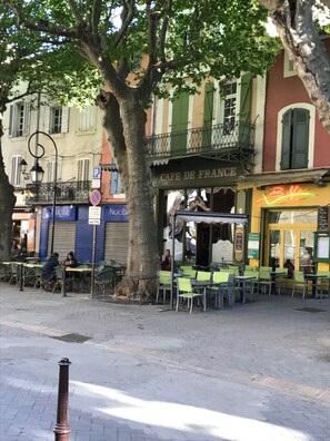 Un café con terraza, un árbol en primer plano y un edificio con balcón al fondo.