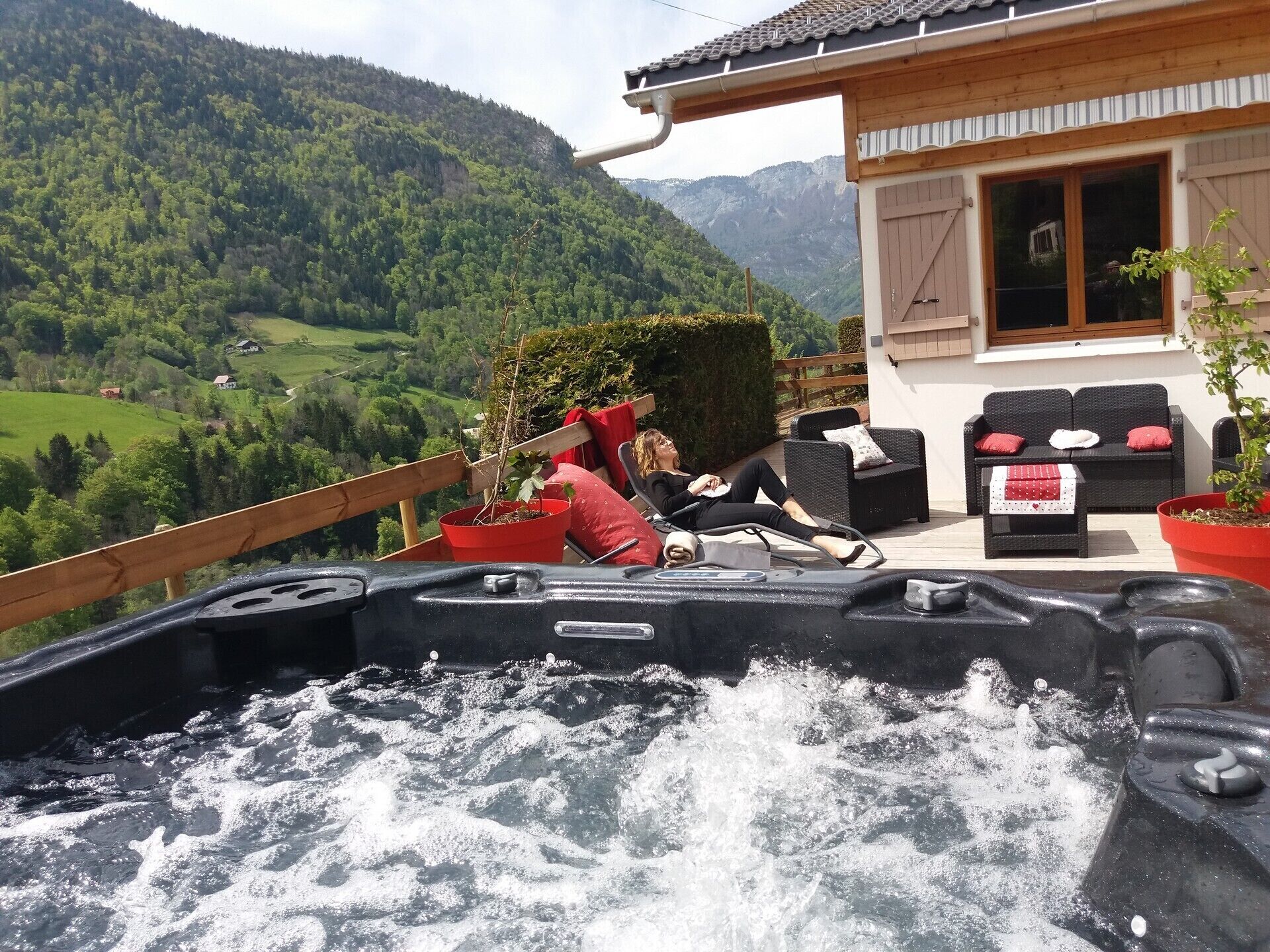 Ein Whirlpool mit einer entspannten Person, umgeben von Gartenmöbeln und Blumentöpfen, mit Blick auf eine Berglandschaft.