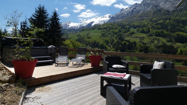 A wooden deck with outdoor furniture, including chairs and a table, overlooking a mountainous landscape with snow-capped peaks and lush greenery. - Chalet Haute Savoie quiet jacuzzi wifi near Lake Annecy (Thônes)