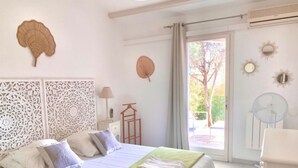 Un dormitorio ordenado con una cama, almohadas, un plaid verde, un panel decorativo en la pared, un ventilador y una ventana con vista a árboles.