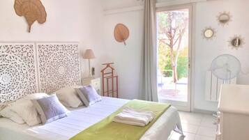 Een netjes opgemaakte slaapkamer met een bed, kussens, een groene dekbedovertrek, een decoratief paneel, een ventilator en een raam met uitzicht op bomen.