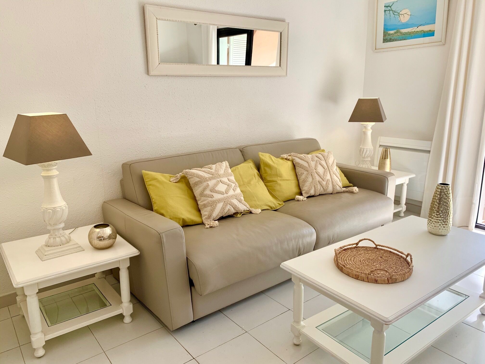 A living room with a beige sofa, yellow and patterned cushions, a white coffee table with a woven tray, a white lamp, and a framed mirror on the wall.