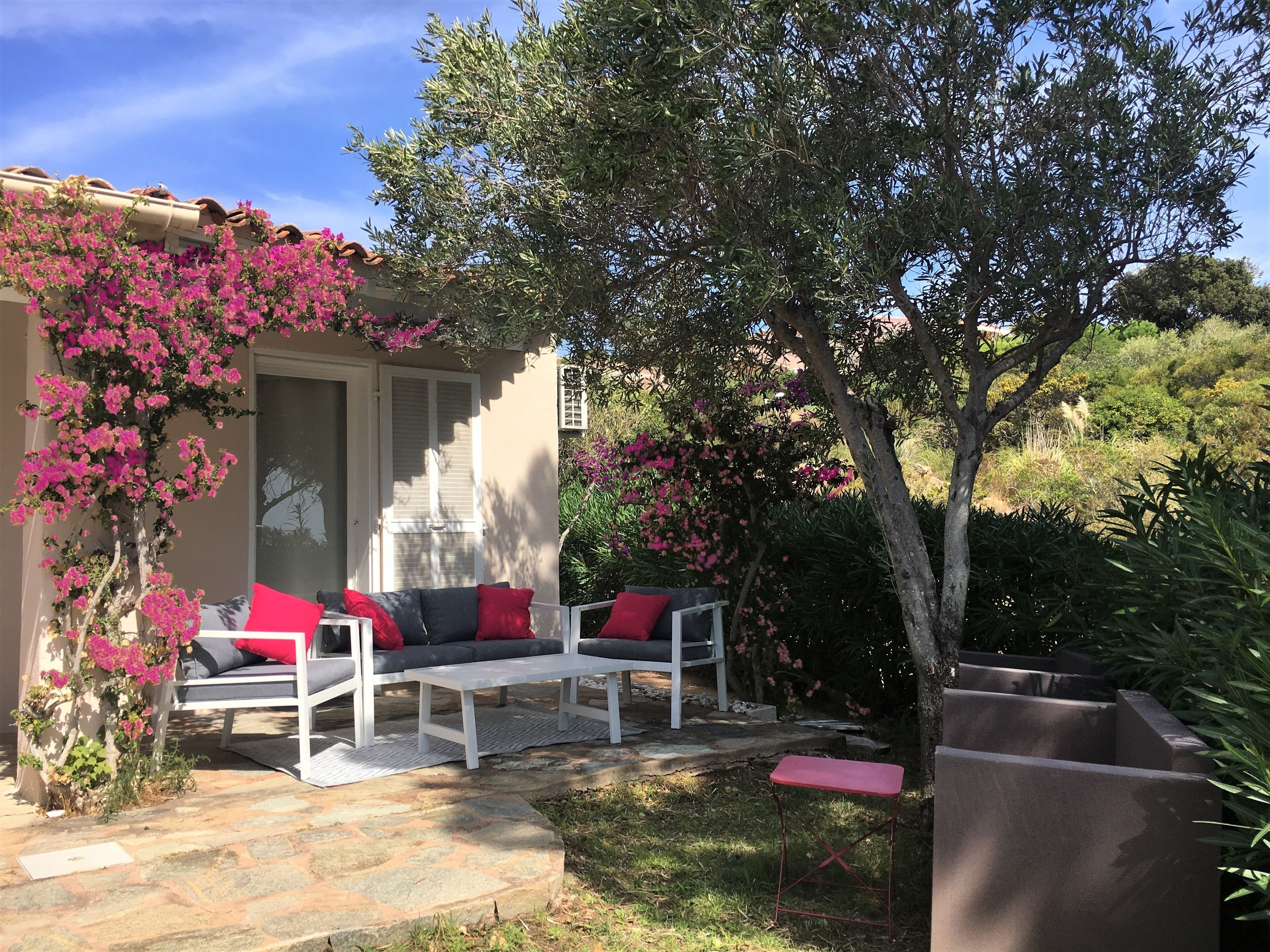 En terrasse med en hvid sofa og et bord, omgivet af lyserøde buganvilleaf blomster, under en klar himmel.