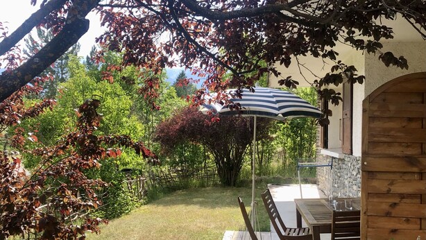 Un patio con mesa y sillas, una sombrilla a rayas y vistas a un jardín con árboles y arbustos.