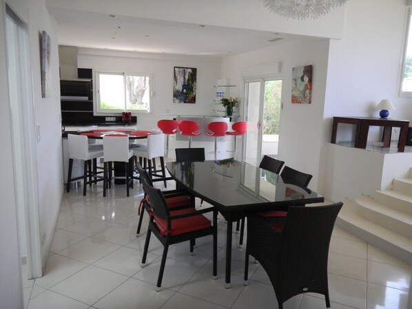 A modern dining area with a glass table and chairs, a kitchen in the background, and artwork on the walls. - Villa, St-Maxime sea view, tranquillity, air-conditioned, heated pool (Sainte-Maxime)