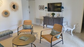 A living room with two wicker chairs, a small round table, a white bench, a dark wooden cabinet, and a flat-screen TV on the wall.