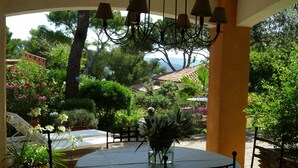 A round table with chairs under a pergola, surrounded by potted plants and a view of a house with a tiled roof.