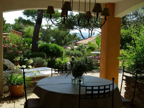 A round table with chairs under a pergola, surrounded by potted plants and a view of a house with a tiled roof.