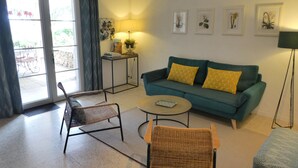 A living room with a sofa, two chairs, a coffee table, and a lamp. There is a window with curtains, a small table with a lamp, and framed pictures on the wall.