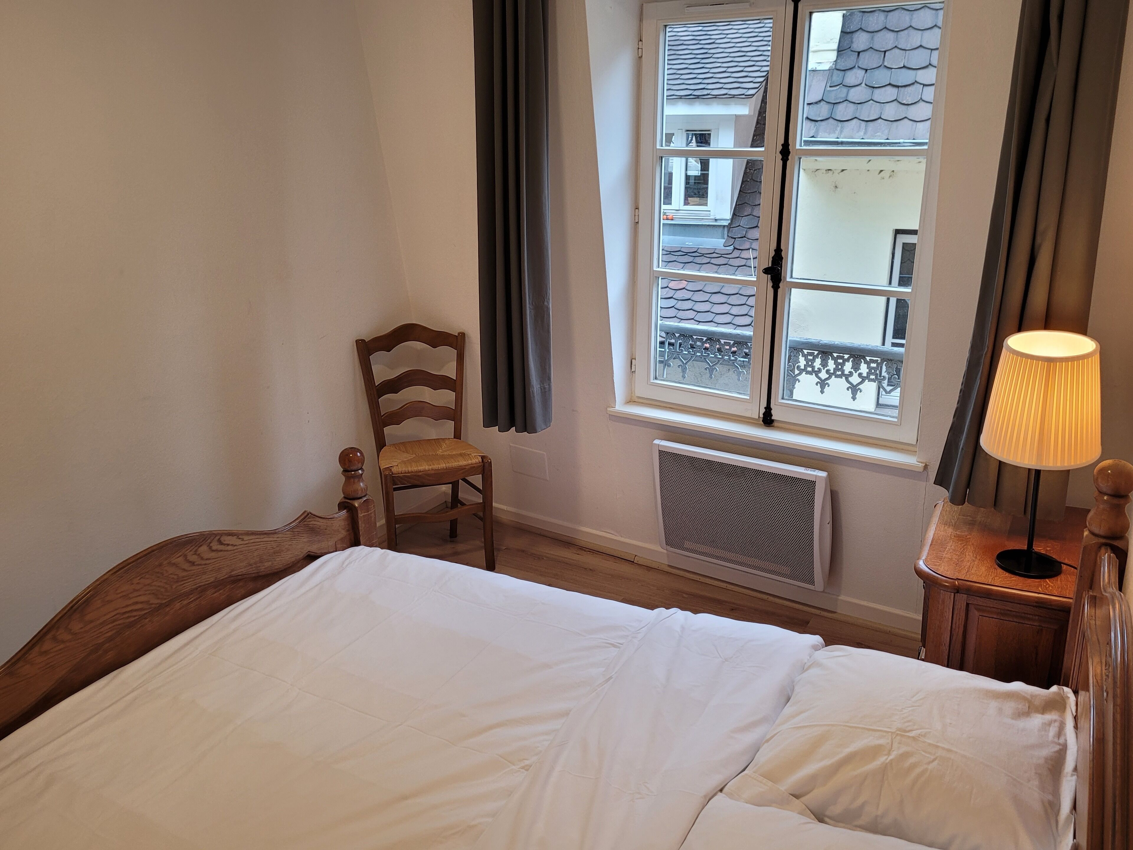 Une chambre à coucher avec un lit, une chaise en bois, une fenêtre avec des rideaux et un radiateur.
